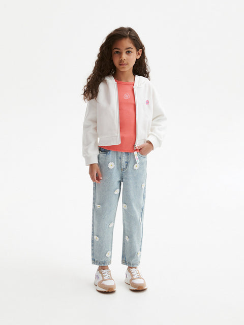 Young girl wearing a white jacket, pink shirt, and light blue jeans with white polka dots on a white background.