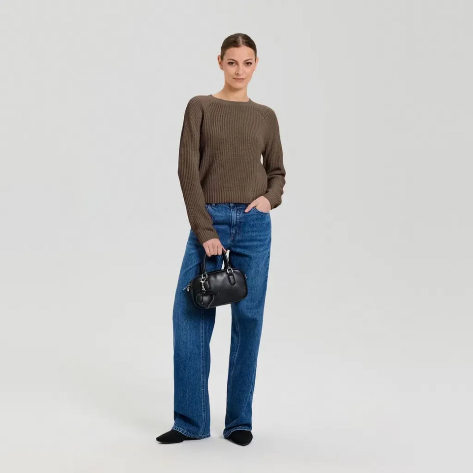 A model stands against a light gray studio background wearing a ribbed brown long-sleeve sweater, blue wide-leg jeans, black socks, and carrying a small black leather handbag.