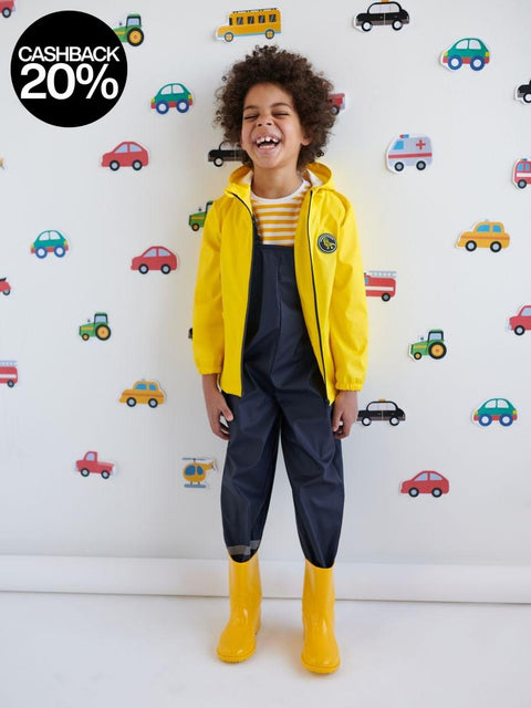 Child wearing a yellow raincoat and boots standing in front of a wall with vehicle pattern