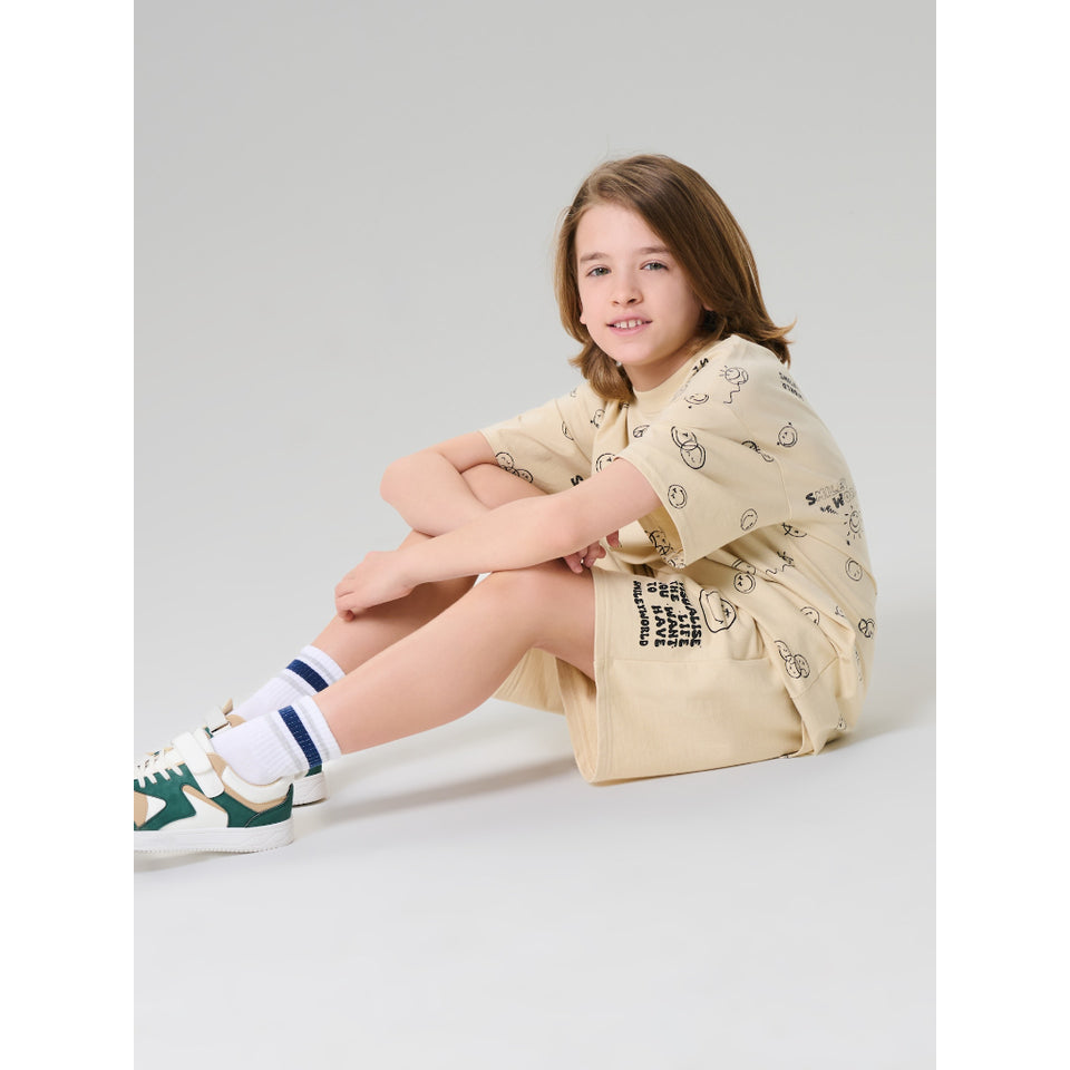 A child model sitting on a light gray studio floor wearing a beige short-sleeve t-shirt dress with black cartoon doodle prints, white socks, and green and white sneakers.
