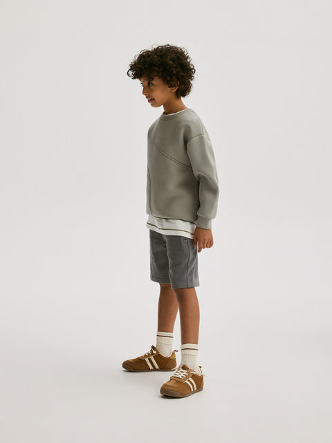 Child wearing a green sweater, gray shorts, and brown shoes on a white background