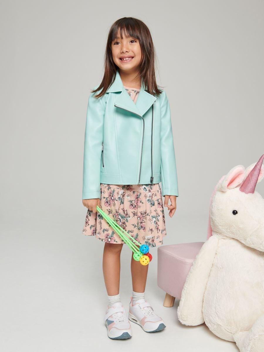 A girl wearing a light blue biker jacket with a classic collar, zip fastening, and two pockets, standing next to a plush unicorn toy.