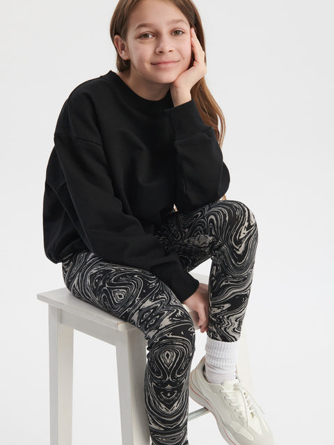 Young girl sitting on a white stool wearing a black sweatshirt and patterned leggings.