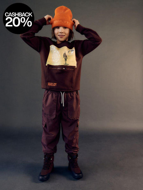 Person wearing a brown sweatshirt with graphic design and matching pants, holding an orange beanie.