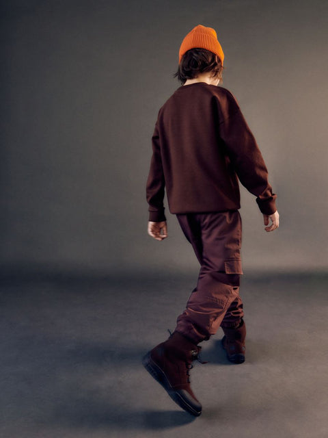 Person wearing a brown outfit with an orange beanie walking on a gray background