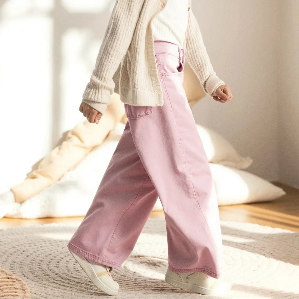 A young girl wearing a cream ribbed cardigan over a white top and pink wide-leg pants, standing in a brightly lit room with sunbeams on the wall.