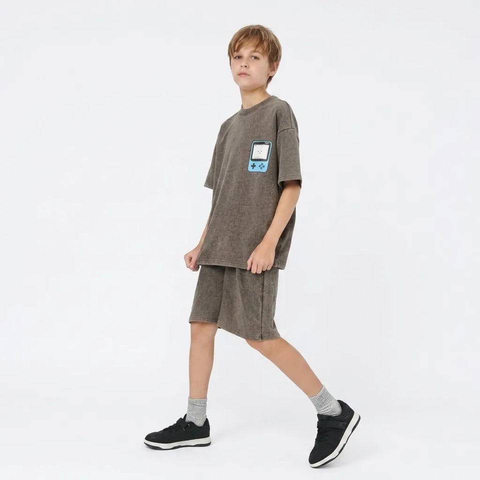 A child model wearing a matching two-piece lounge set consisting of an oversized short-sleeve t-shirt and shorts in a heathered taupe-brown color. The t-shirt features a small blue and white retro game console graphic on the left chest.