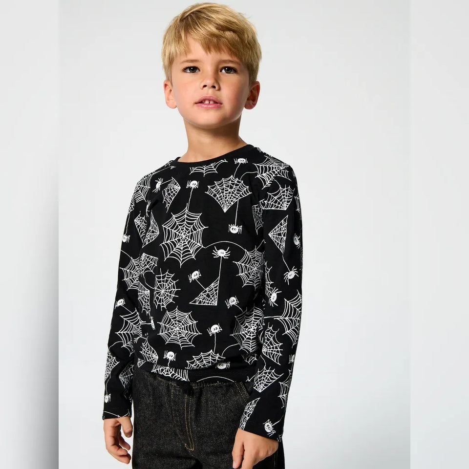 A young boy with blonde hair wearing a black long-sleeve crewneck shirt with a white spiderweb and spider print, paired with dark gray jeans, standing against a light gray studio background.