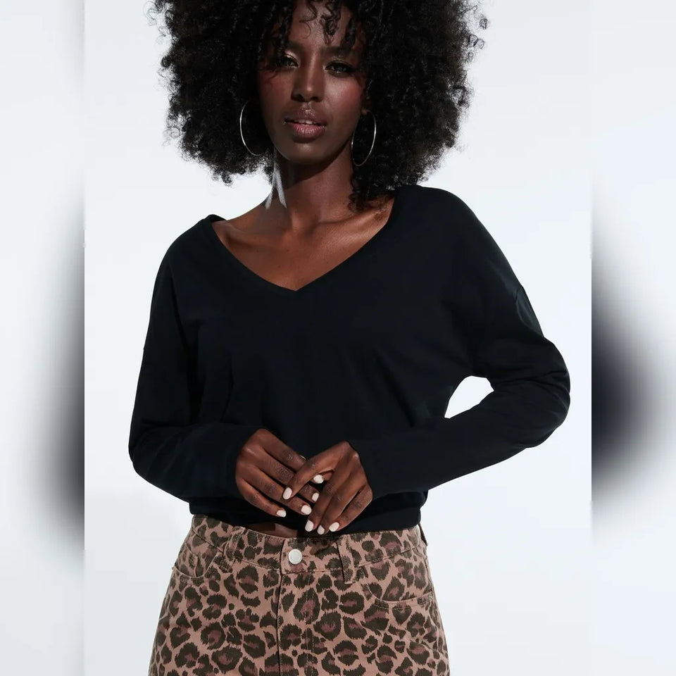A woman with a large black afro wearing a black long-sleeve V-neck top and leopard print pants, standing against a white studio background.