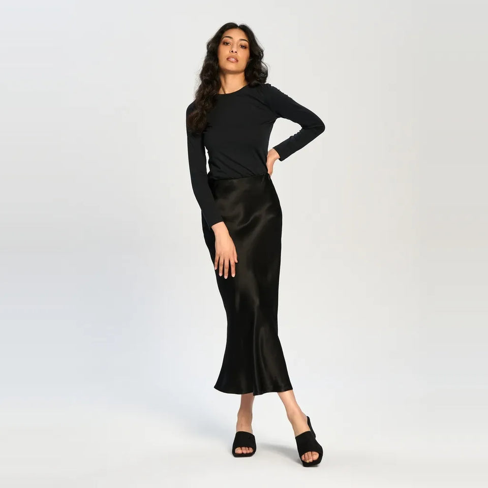 A woman modeling a black long-sleeve knit top and a black satin midi skirt, paired with black open-toe heeled sandals, against a plain light gray studio background.