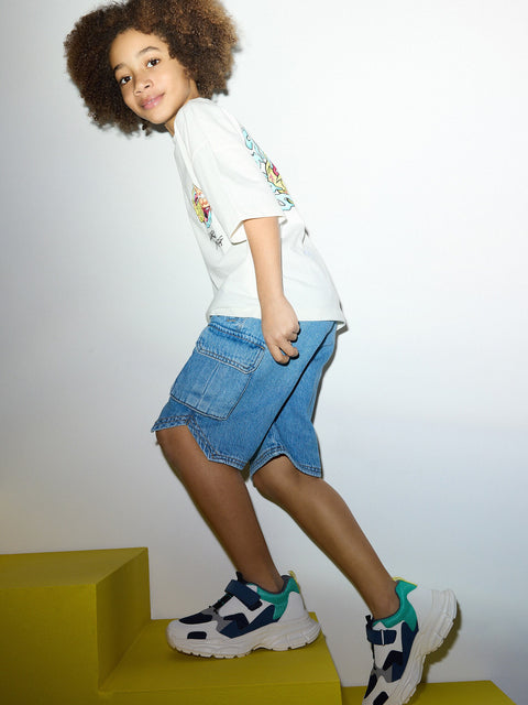 Child wearing a white t-shirt with colorful graphics, blue denim skirt, and white sneakers with blue accents on a plain background.
