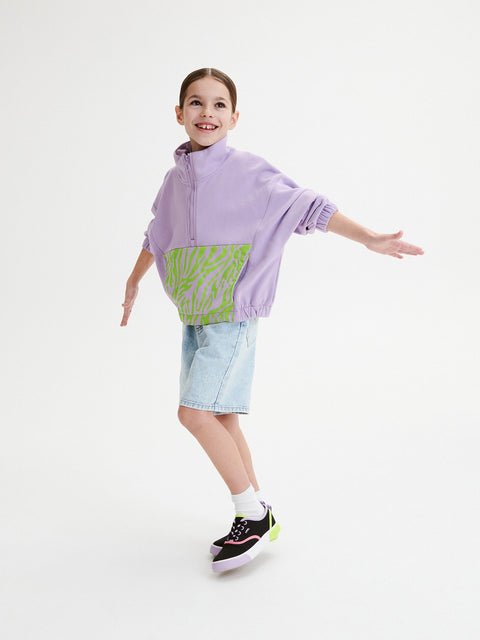 Child wearing a purple poncho with green zebra print and denim shorts on a white background