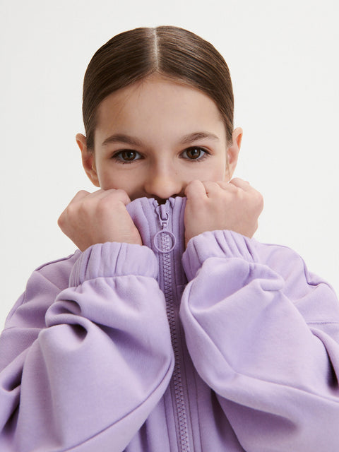 Child wearing a purple jacket with hands covering mouth on a light gray background