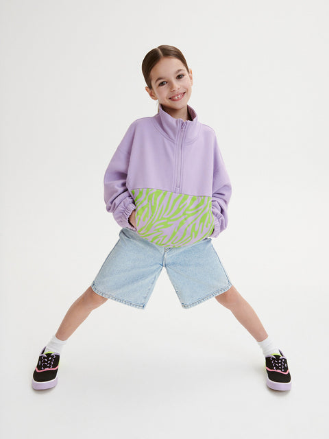 Child wearing a purple jacket with a colorful pattern, denim shorts, and sneakers on a white background
