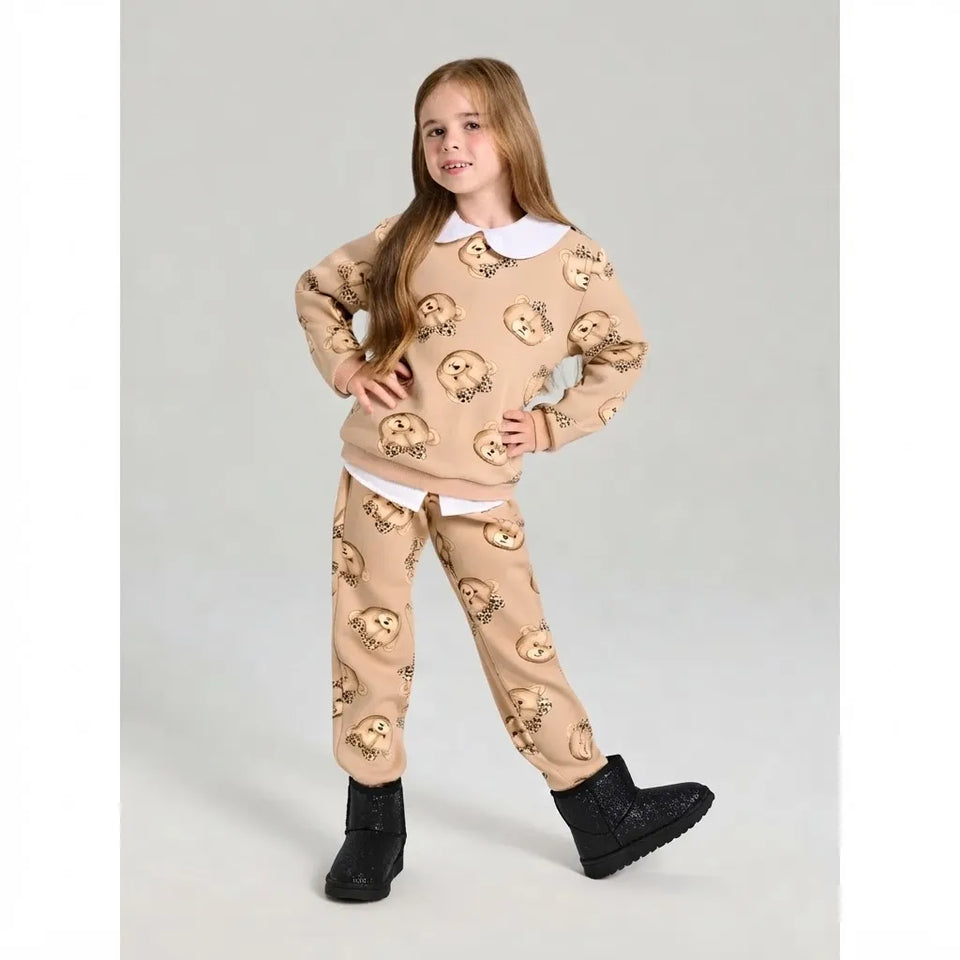 A young girl modeling a beige two-piece pajama set with an owl pattern, featuring a long-sleeve top with a white collar and matching pants, paired with black boots, against a plain light gray background.
