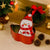 A red metal Christmas ornament holder featuring a painted snowman character wearing a red hat and scarf, styled on a beige fabric surface with pinecones, a wreath, and holiday decorations in the background.