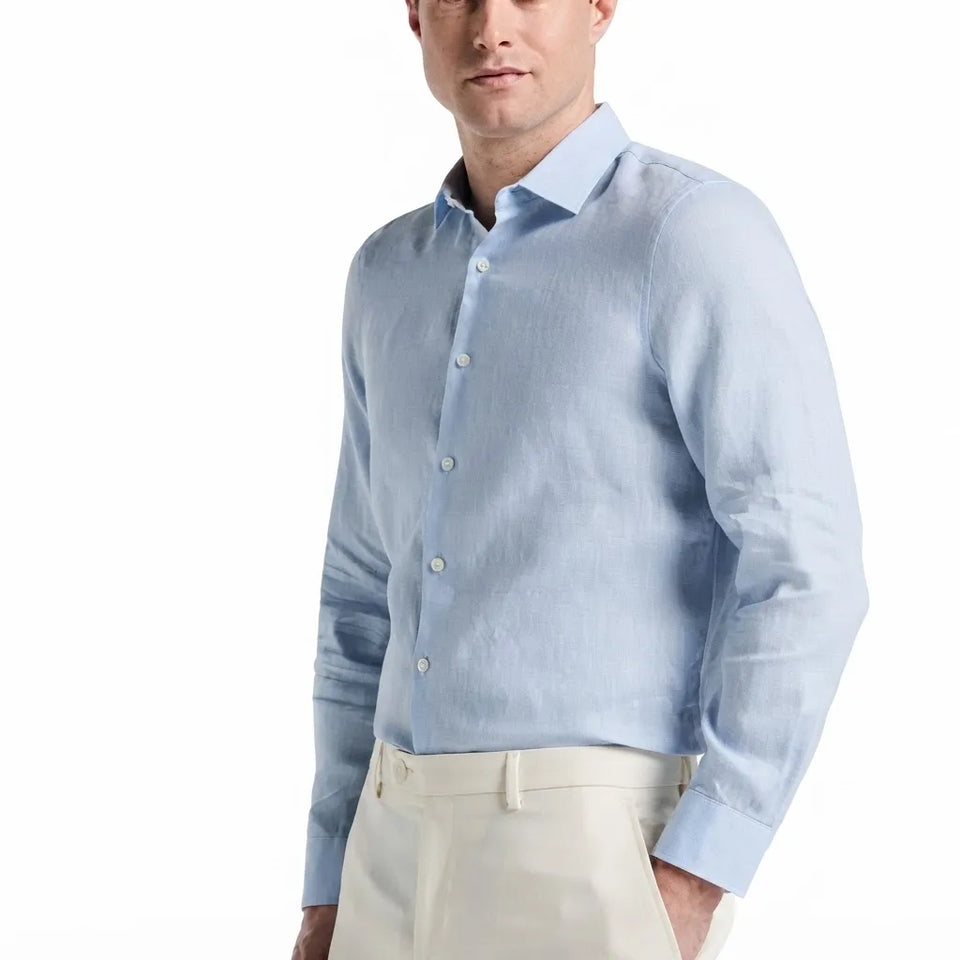 A man wearing a light blue long-sleeve linen button-down shirt and cream trousers, shown from the chest up against a plain white background.