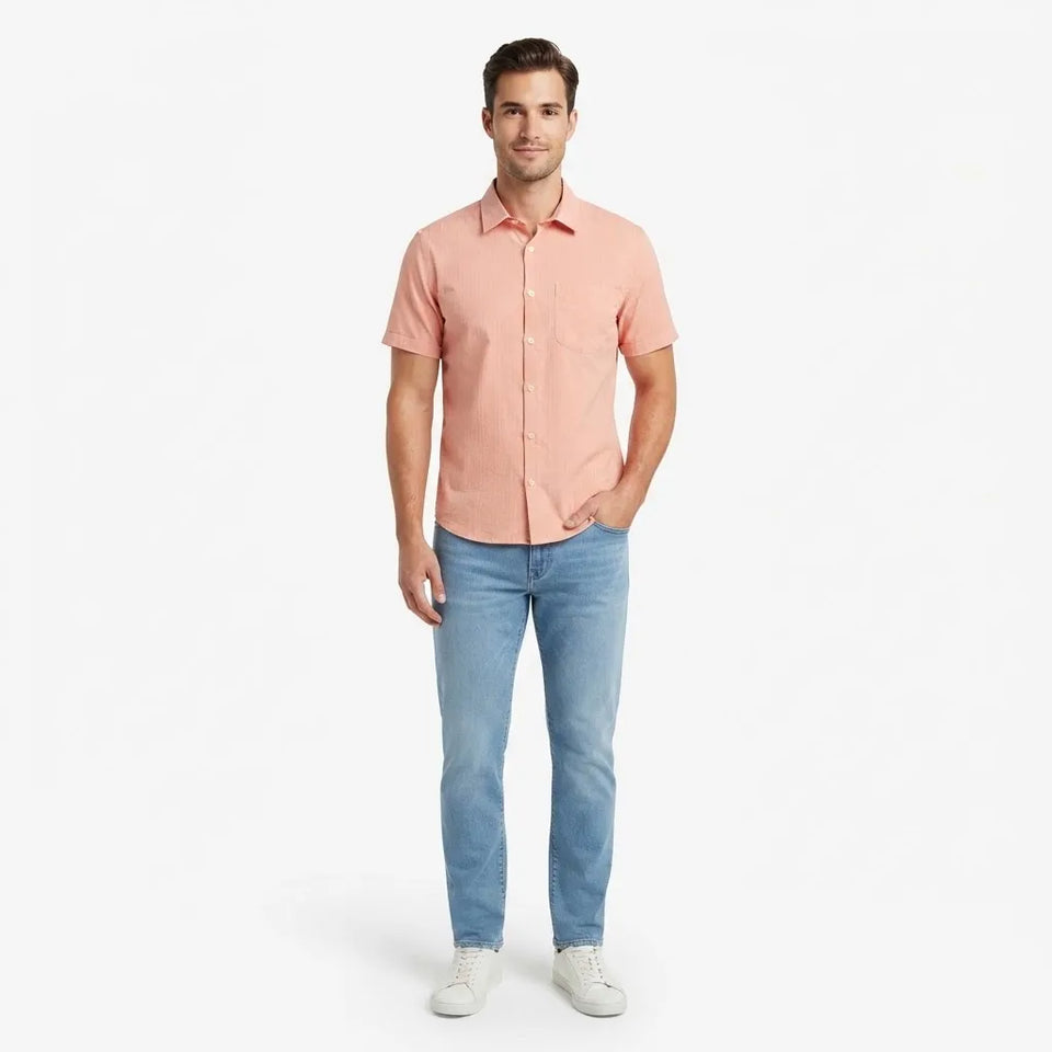 A man modeling a short-sleeve button-up shirt in a dusty coral pink color, paired with light blue jeans and white sneakers, against a plain white studio background.