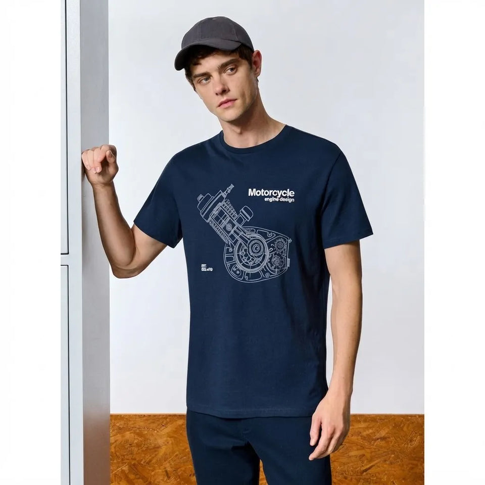 A man wearing a navy blue crewneck t-shirt with a white motorcycle engine schematic graphic on the front, paired with a dark gray baseball cap and matching navy pants, standing against a white wall with a wood floor.