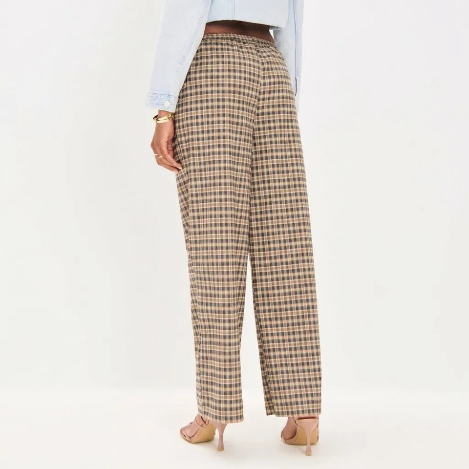 A person shown from the back wearing beige plaid wide-leg trousers with a brown belt, a light blue shirt, and beige heels against a plain white studio background.