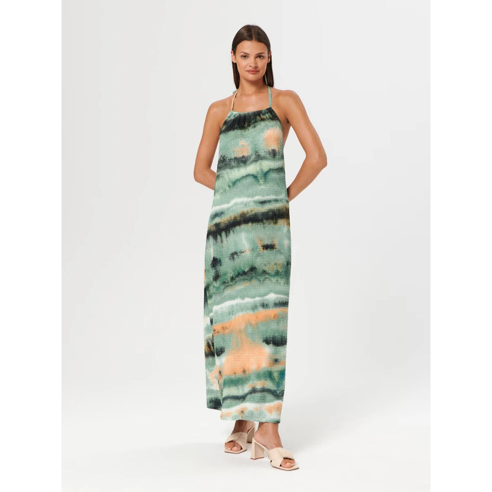 A woman modeling a sleeveless halter-neck maxi dress with an abstract watercolor-style print in shades of teal, green, black, and peach, paired with beige heeled sandals against a light gray studio background.