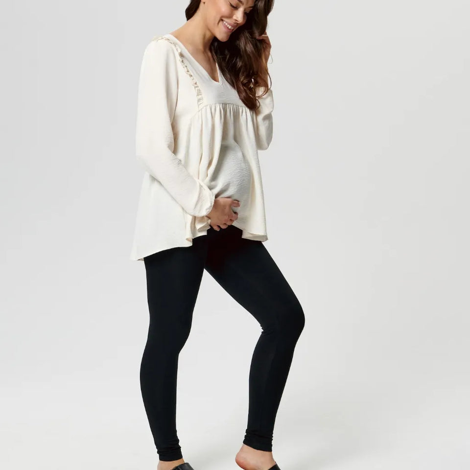 A pregnant woman wearing a cream-colored long-sleeve top with decorative lace trim and black leggings, standing against a light gray studio background.