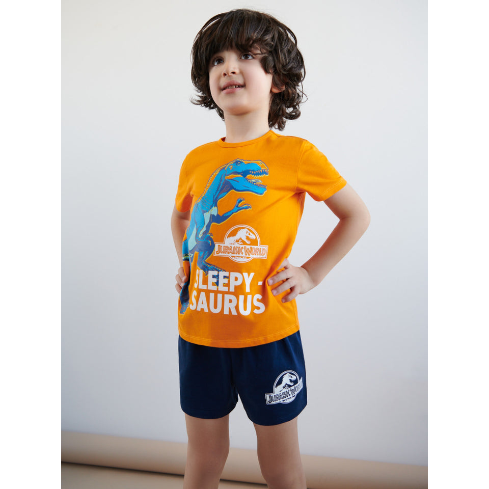 A young boy modeling a matching Jurassic World pajama set, featuring a bright orange t-shirt with a blue T-Rex graphic and 'Sleepy Saurus' text, paired with navy blue shorts.