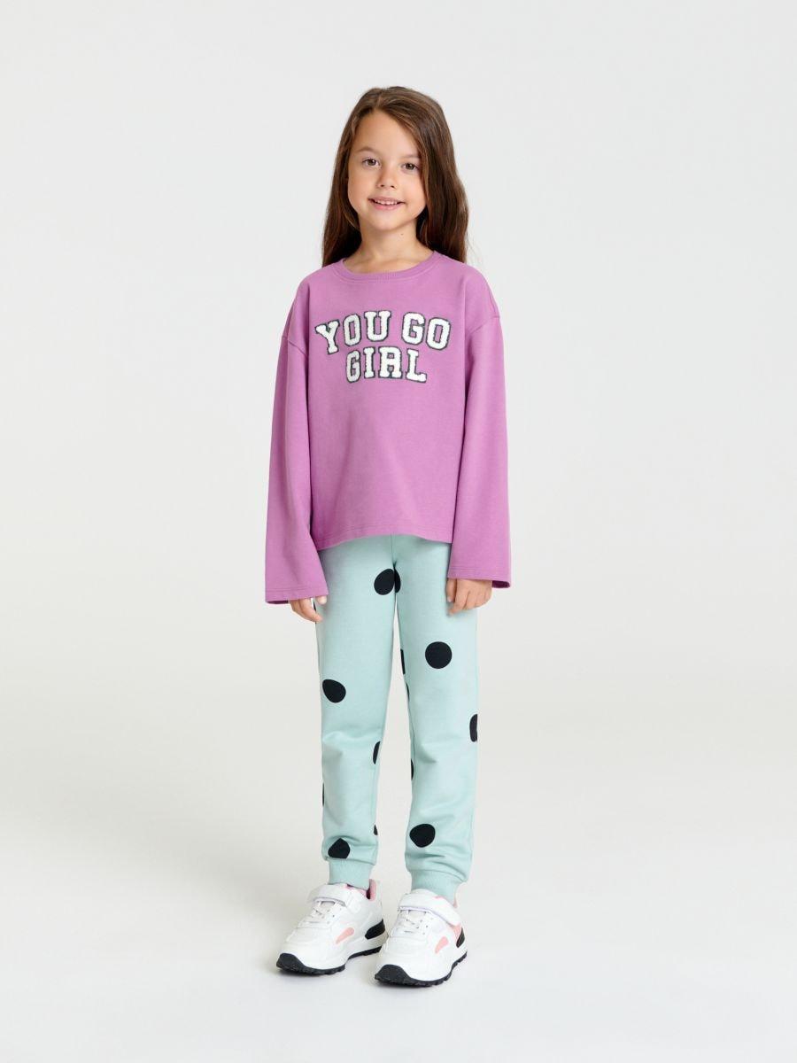 Young girl wearing a pink sweatshirt with 'YOU GO GIRL' text and light blue pants with black polka dots on a white background.