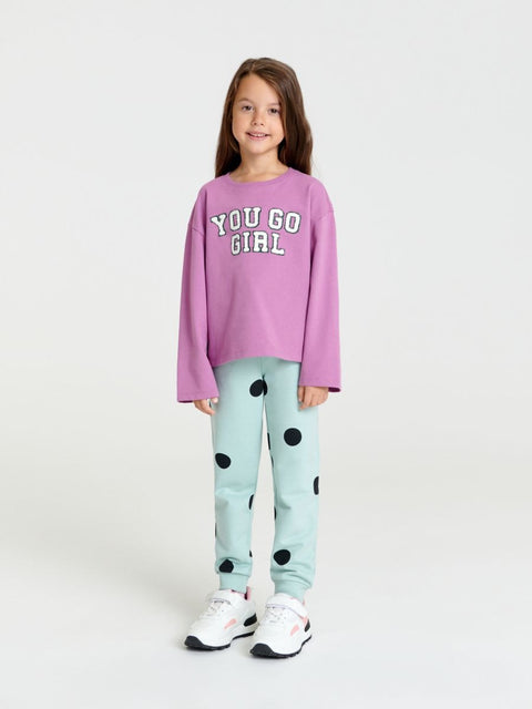 Young girl wearing a pink sweatshirt with 'YOU GO GIRL' text and light blue pants with black polka dots on a white background.
