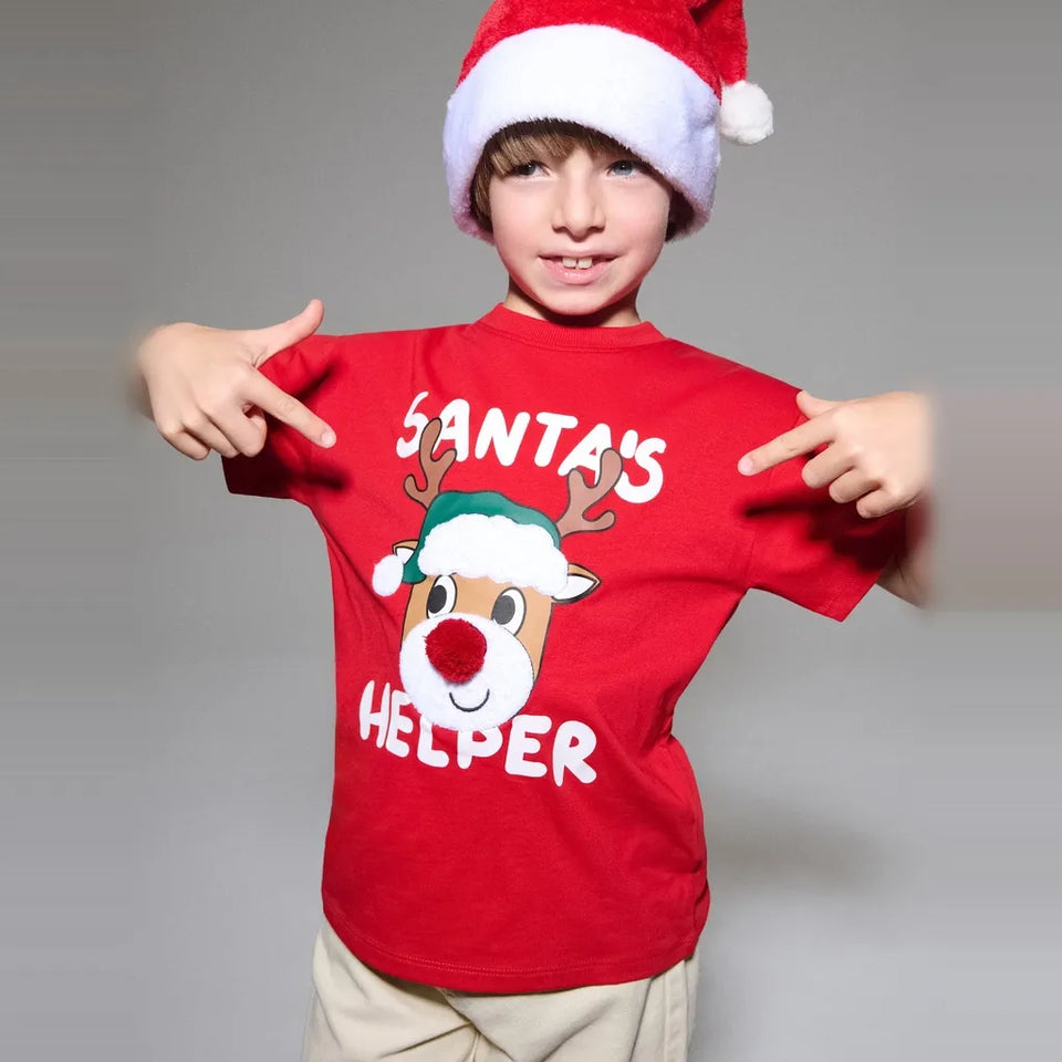 A young boy wearing a red Santa hat and a red t-shirt with a cartoon reindeer and the words 'SANTA'S HELPER', pointing at the shirt against a plain gray background.
