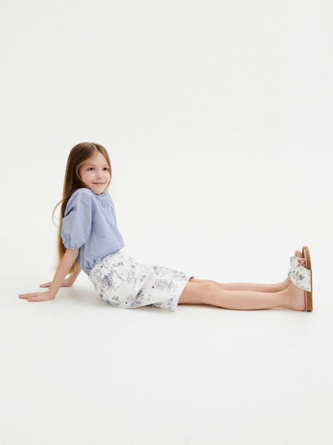 Young girl sitting on a white floor wearing a blue top and floral skirt.