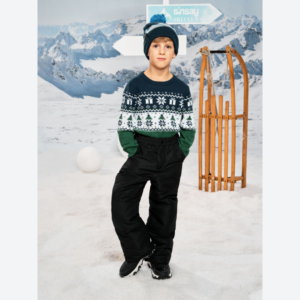 A child wearing a navy and green knit sweater with a white Nordic pattern, black snow pants, and a blue pom-pom beanie stands in a snowy mountain setting with a wooden sled and a sign that reads 'insay SKI CLUB'.