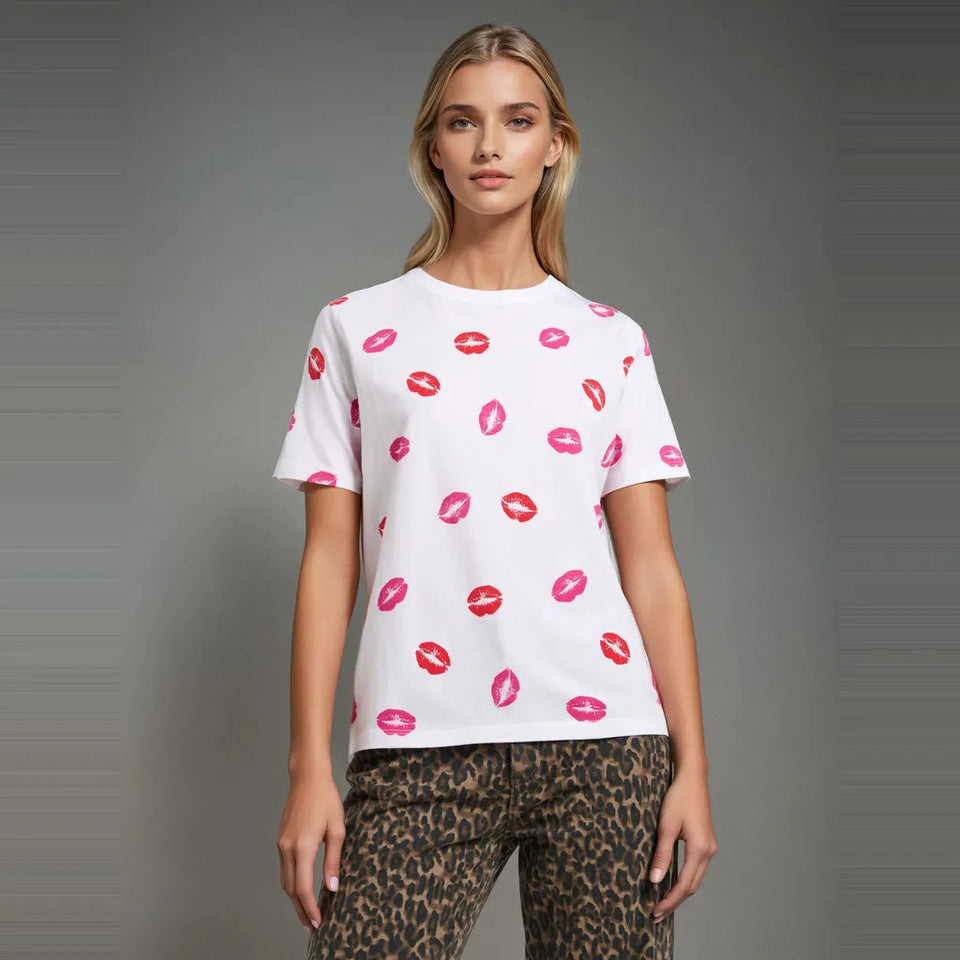 A woman modeling a white short-sleeve crewneck t-shirt with an allover pink and red lipstick kiss print, paired with leopard-print pants, against a neutral gray studio background.