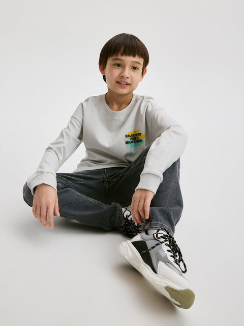 Child sitting on the floor wearing a light gray sweatshirt with a colorful graphic and dark pants.
