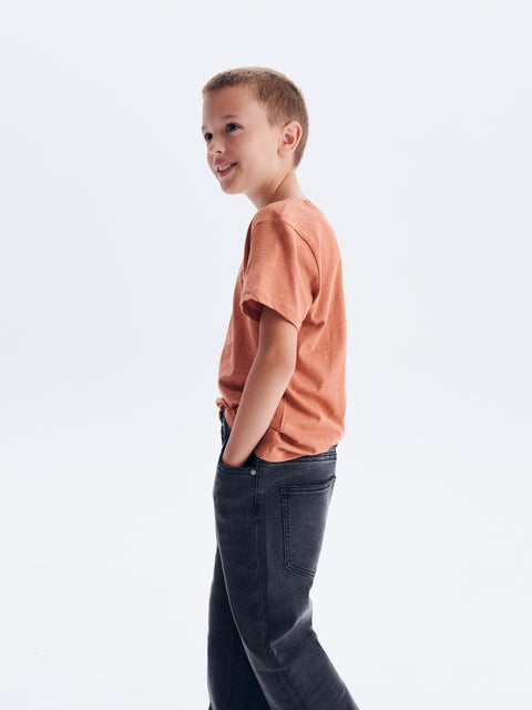 Young boy wearing a rust-colored t-shirt and dark jeans on a white background