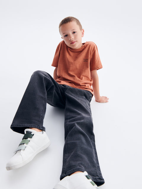 Child wearing a brown t-shirt, dark jeans, and white sneakers on a light gray background