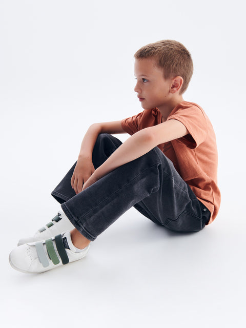 Young boy sitting on a white floor wearing a brown shirt and dark pants.