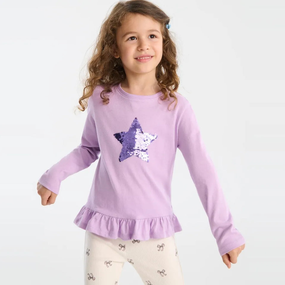 A young girl with curly brown hair wearing a lavender long-sleeve top with a purple and white sequined star on the chest, paired with cream patterned leggings, against a plain white background.