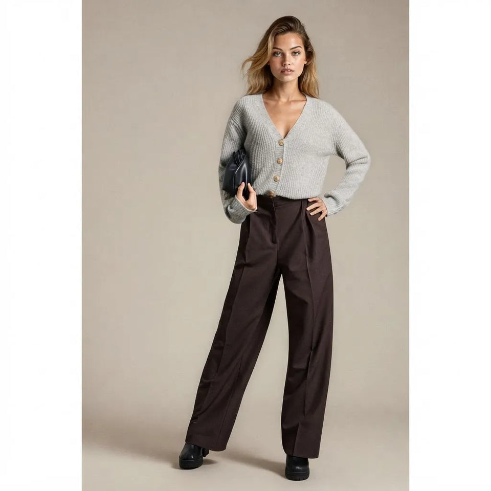A model stands against a plain beige studio background wearing a light gray knit cardigan with gold buttons, dark brown wide-leg trousers, and black shoes, while holding a dark clutch.