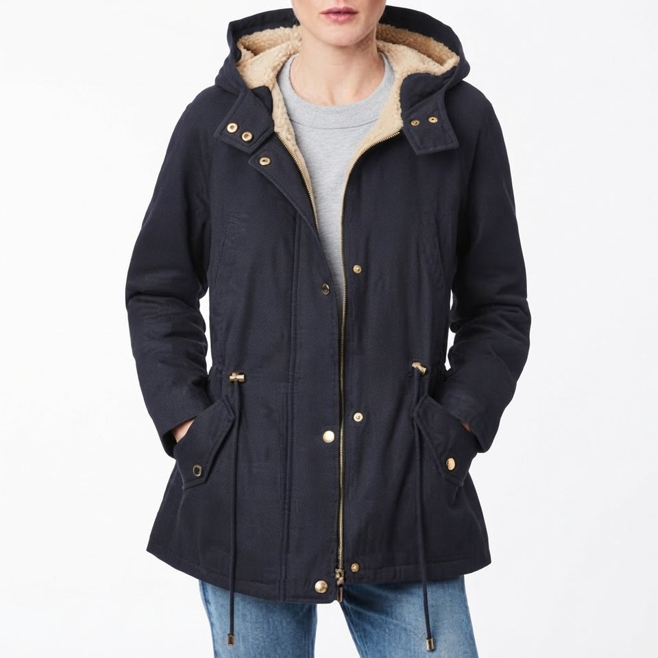 A woman wearing a dark navy hooded jacket with a beige sherpa lining, gold-tone snap and zipper details, and front patch pockets, paired with a gray shirt and blue jeans against a plain white background.