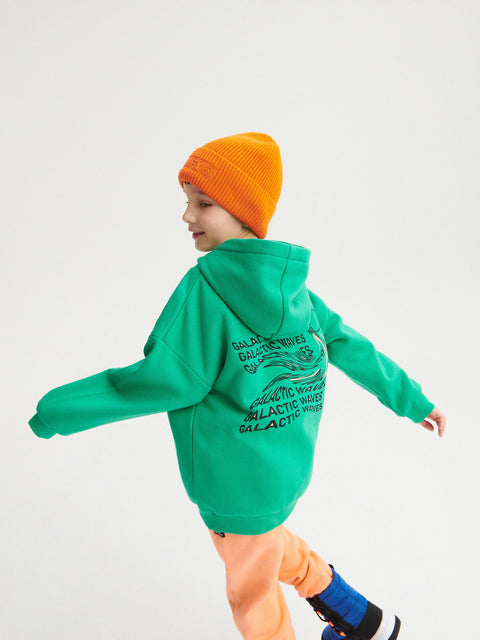 Child wearing a green hoodie with text, orange pants, and an orange beanie on a light gray background