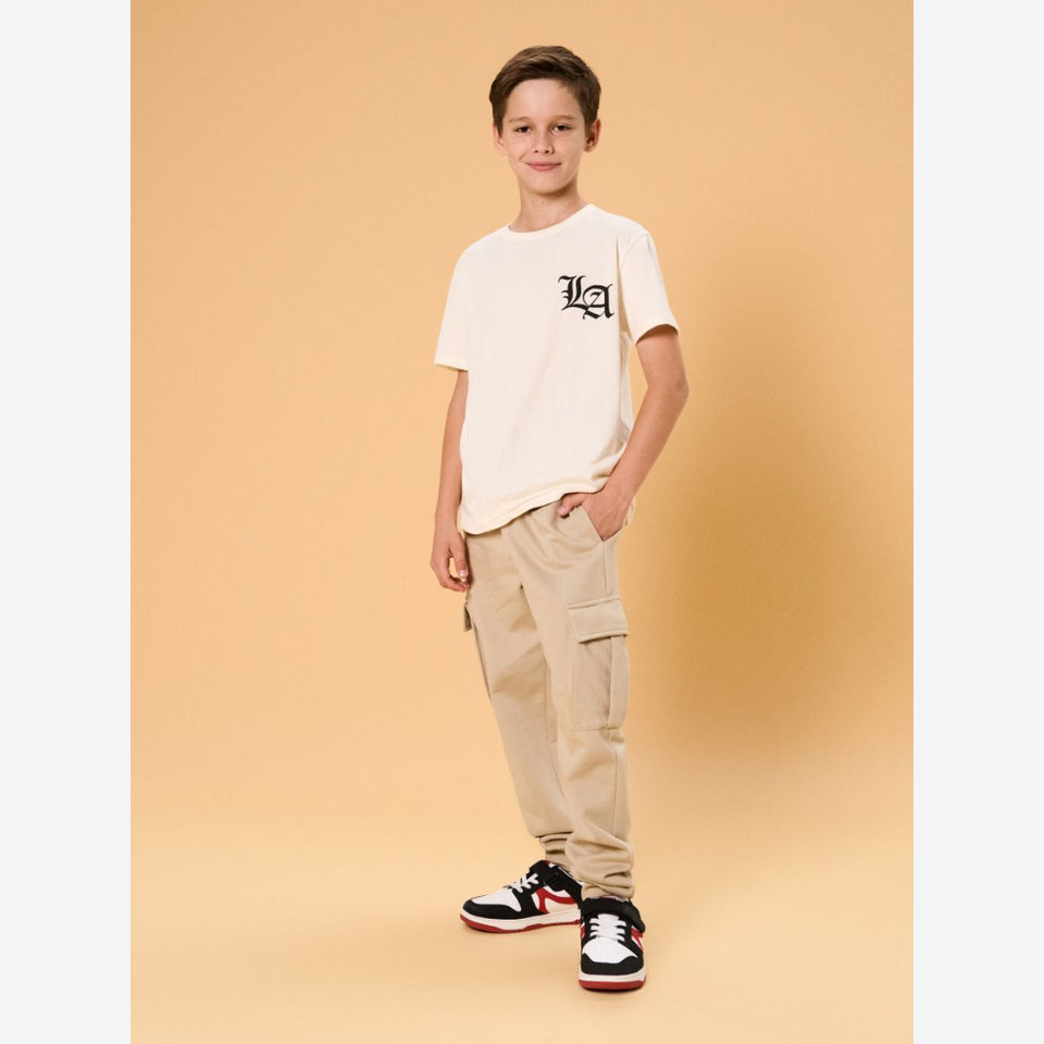 A young boy stands against a solid beige studio background wearing a cream-colored t-shirt with a black graphic logo on the chest, beige cargo pants, and black and white sneakers with red accents.