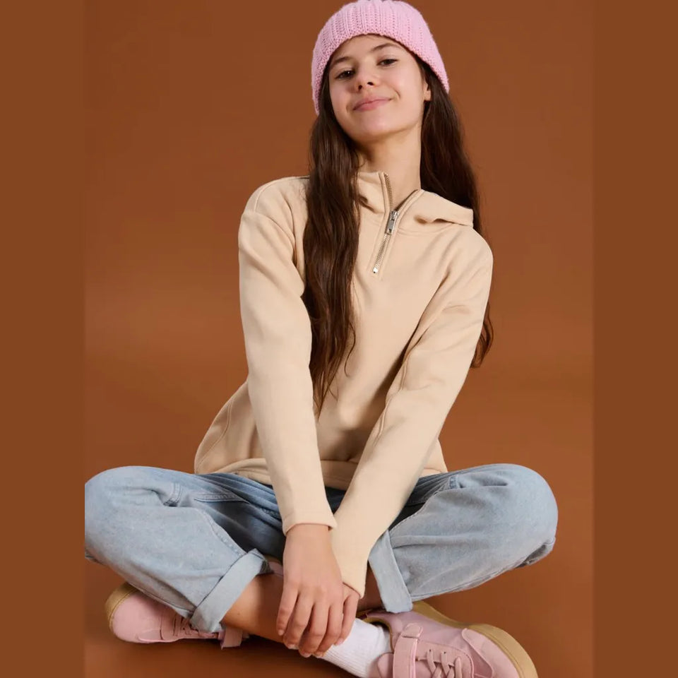 A young girl sitting cross-legged against a solid brown background, wearing a beige half-zip hoodie, light blue jeans, a pink beanie, and pink sneakers.