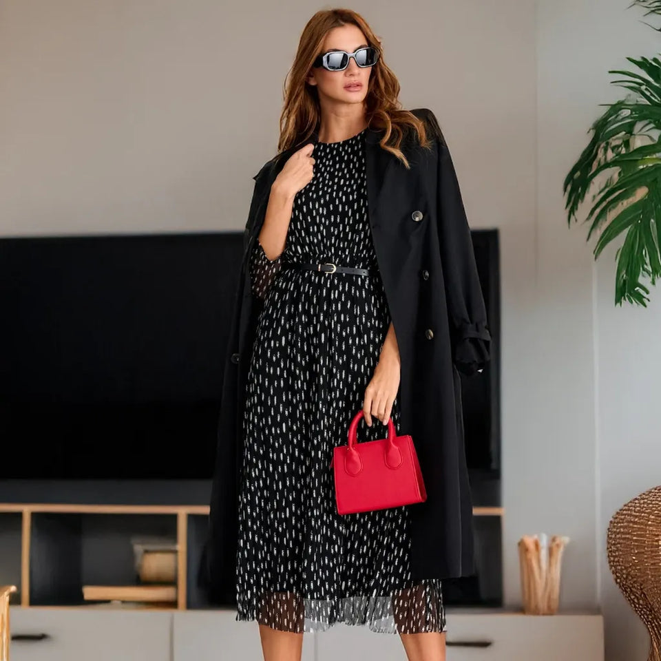 A woman models a black long coat over a black midi dress with a white geometric pattern, accessorized with a red handbag and sunglasses in a modern living room setting.