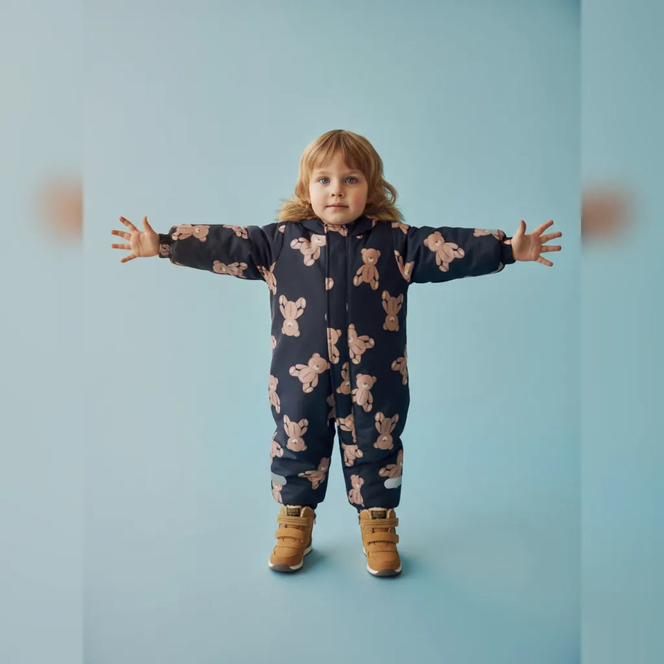 A young child stands with arms outstretched wearing a dark navy one-piece snowsuit covered in a light pink teddy bear print, paired with tan boots, against a solid light blue background.