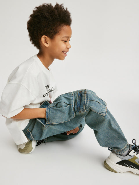 Child wearing a white shirt, blue jeans, and black and white sneakers on a light gray background
