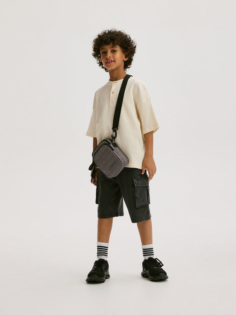 Child wearing a white shirt, black cargo shorts, and carrying a gray bag on a white background