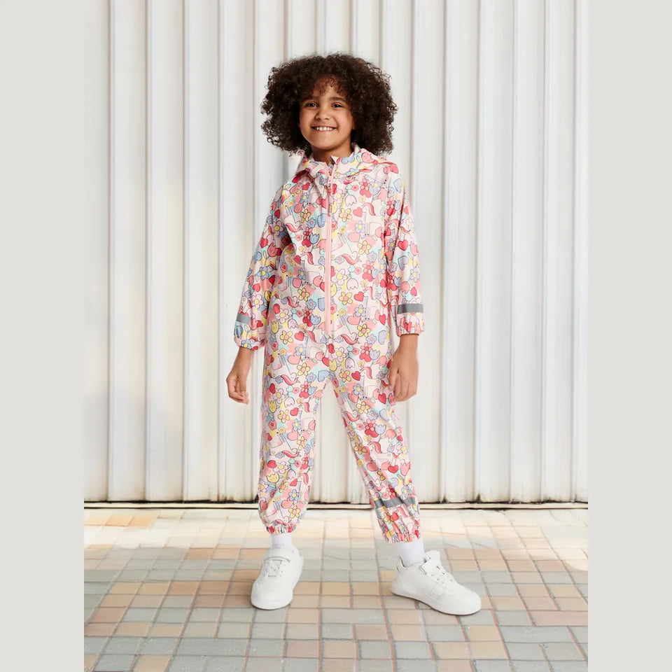 A young child wearing a hooded one-piece jumpsuit with a colorful retro bubble print, standing on a tiled floor in front of a white corrugated metal wall.