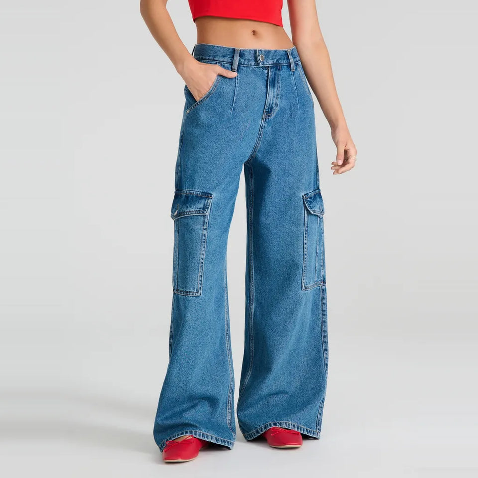 A model wearing high-waisted, wide-leg blue denim cargo pants with large patch pockets on the thighs, styled with a red crop top and red shoes against a light gray studio background.