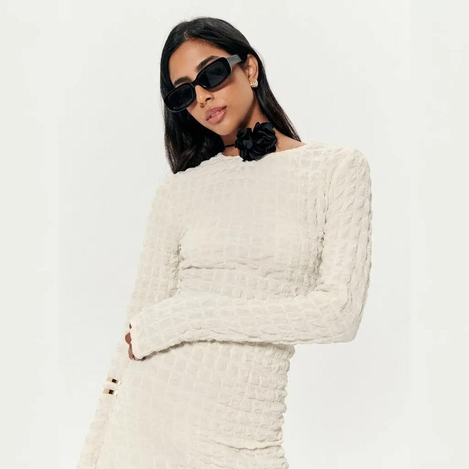A woman wearing a textured off-white long-sleeve dress with a black fabric flower at the neckline, styled with black sunglasses and a black flower accessory, against a plain white background.